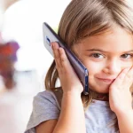 Ein junges Mädchen hält ein HTC-Smartphone an ihr Ohr und schaut konzentriert nach vorne. Im Hintergrund ist ein unscharfer neutraler Hintergrund zu erkennen.